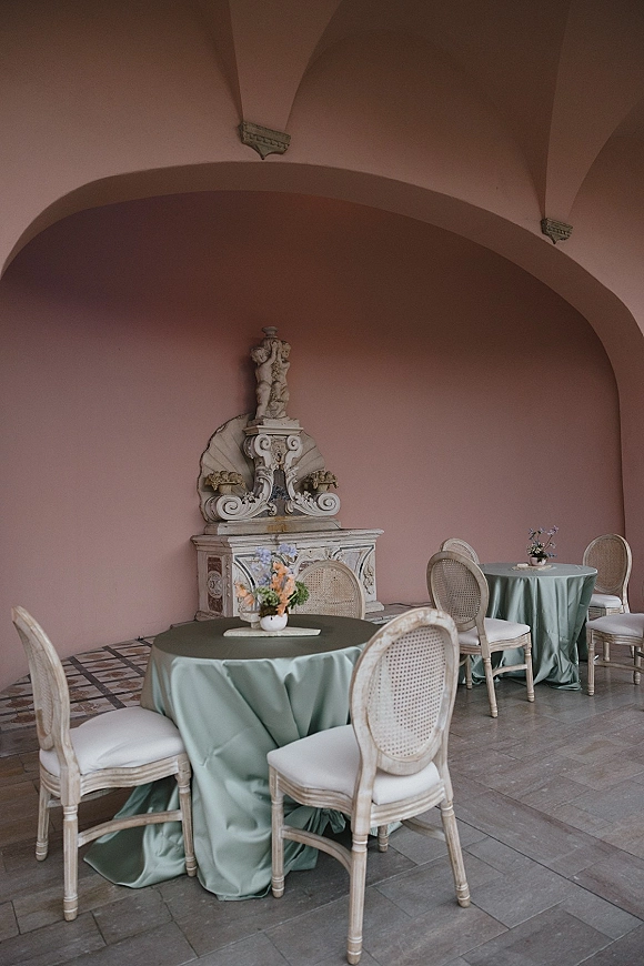 Wedding reception tablescape with round wedding reception tables in sage green linens, floral centerpieces and rattan chairs by a stone fountain in a pink courtyard