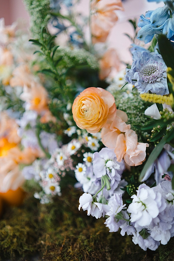 Wedding flowers in a pastel wedding flowers arrangement with peach ranunculus accents, blue blooms, white daisies, and greenery on moss, blurred florals behind