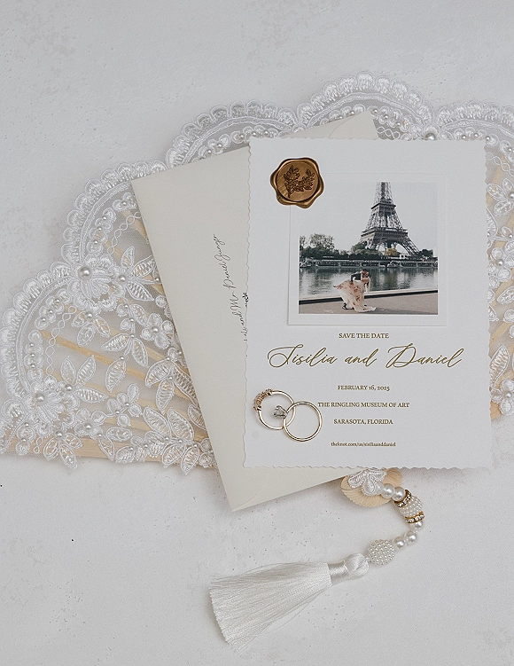 Save the date card with wedding rings, wax seal, and photo insert beside an envelope on lace fabric with pearl details on a white surface