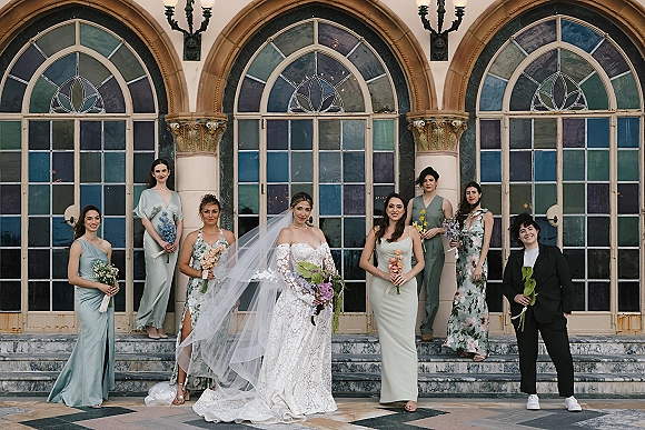 Wedding party portrait with bride and bridesmaids posing with bouquets on stone steps before arched stained glass windows, long veil accent
