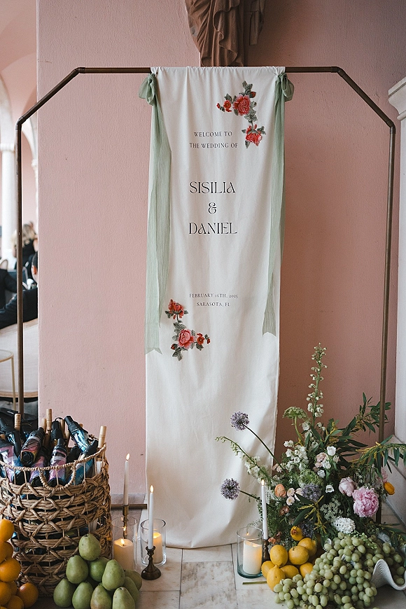 Wedding welcome sign with sage green fabric draping and embroidered florals, styled with candles, greenery, wine and fruit against a pink wall