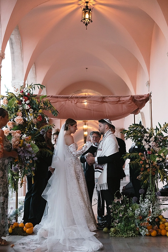 Wedding ceremony moment during a Jewish wedding ceremony as bride and groom exchange vows under a chuppah in an arched corridor with lantern lights