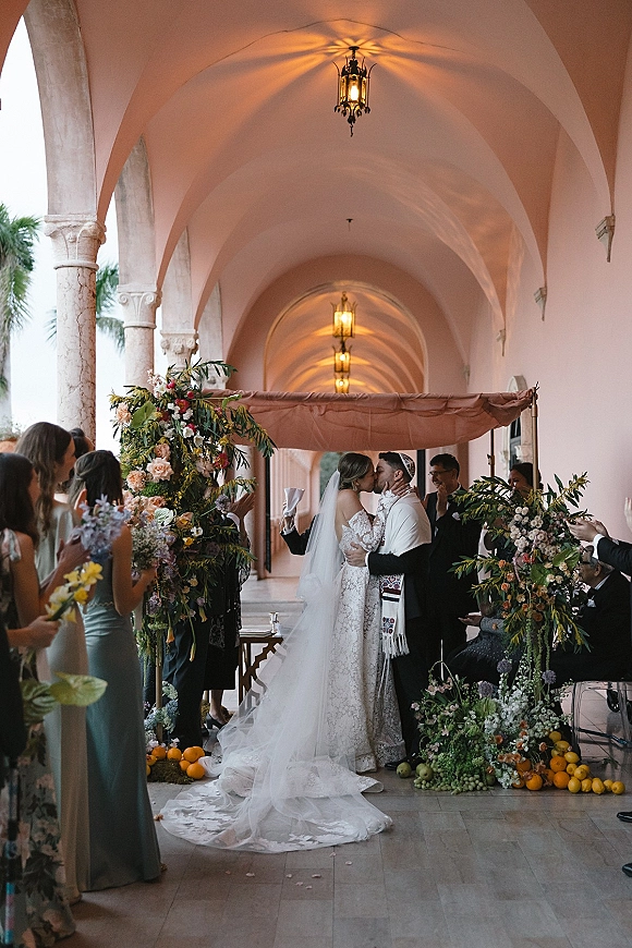 Wedding kiss under a chuppah ceremony as guests cheer, bride in lace veil and groom in tallit amid florals in an arched lantern corridor