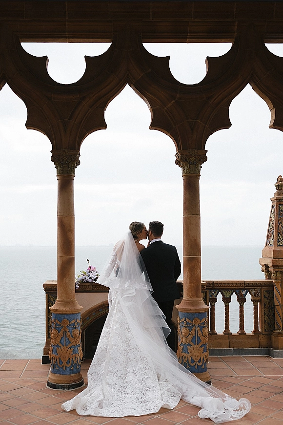 Wedding kiss portrait of bride and groom kiss from behind on a seaside balcony, lace gown and veil with bouquet under ornate archway