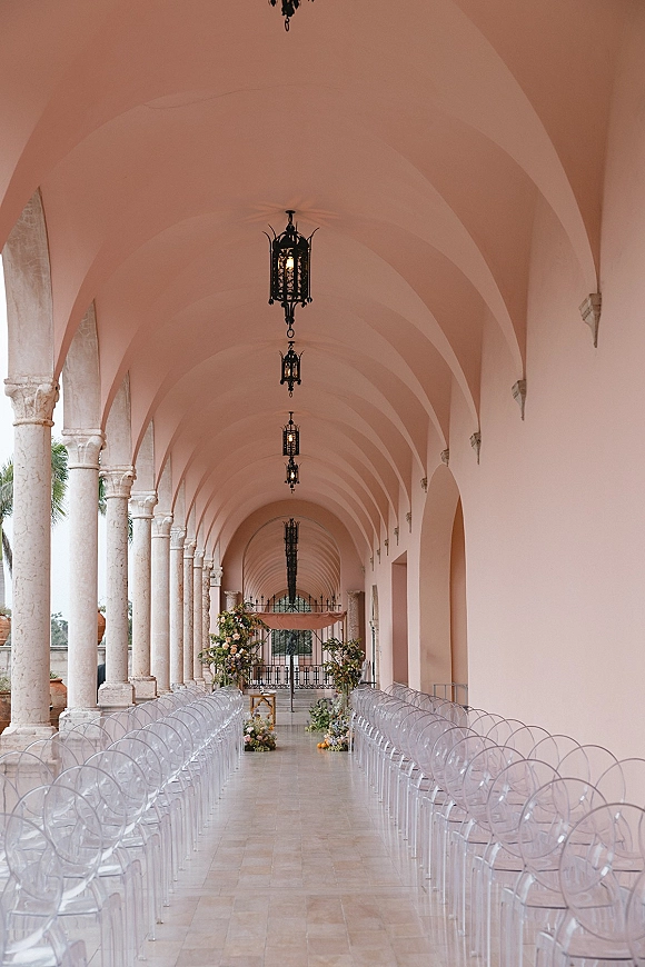 Ceremony setup with clear acrylic chairs lining a long aisle, floral clusters and draped arch beneath hanging lanterns in an arched corridor