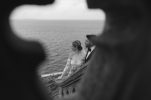 Couple portrait, black and white wedding portrait of bride in lace sleeves and veil with groom in tuxedo by an ocean rooftop terrace railing