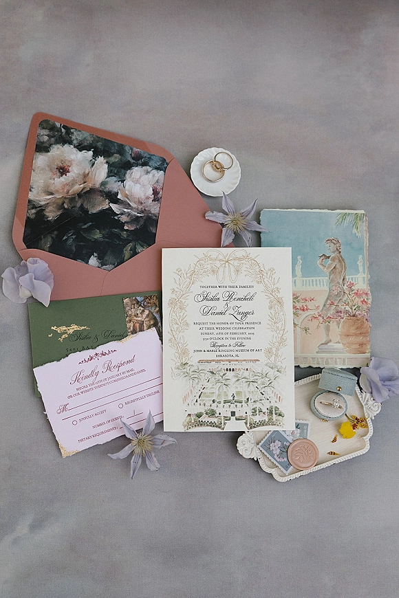 Invitation suite wedding invitation flat lay with wax seal, postage stamp, rings and ribbon beside flowers on a gray fabric backdrop