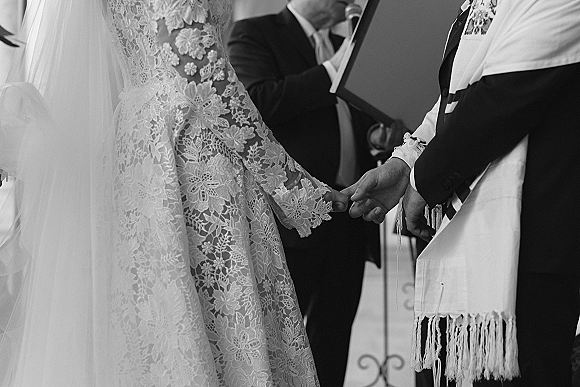 Wedding vows as bride and groom hold hands, lace sleeve and tallit visible beside a microphone stand in a neutral indoor space