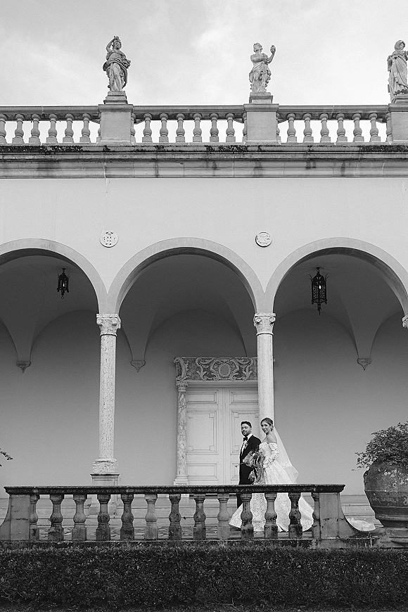 Couple portrait in a black and white wedding portrait, bride in strapless lace gown with bouquet beside groom in tux on colonnade balcony