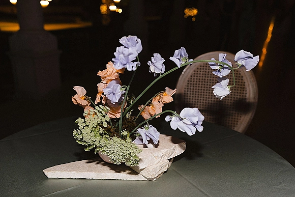 Wedding centerpiece in a stone bowl centerpiece with pastel sweet peas and lace flower on a dining table under warm bistro lights