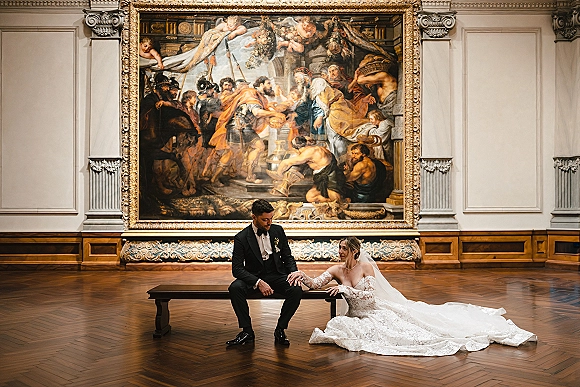 Couple portrait in a museum wedding portrait setting, bride in lace sleeves and long veil beside groom in tux on a bench by a grand framed painting