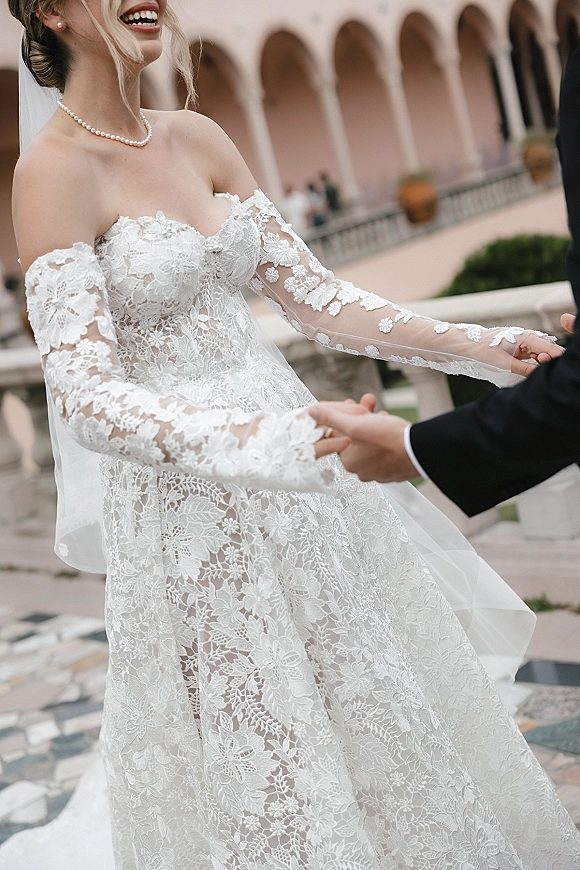 Couple portrait of bride and groom holding hands, bride laughing in lace wedding dress with cathedral veil under an arched colonnade courtyard