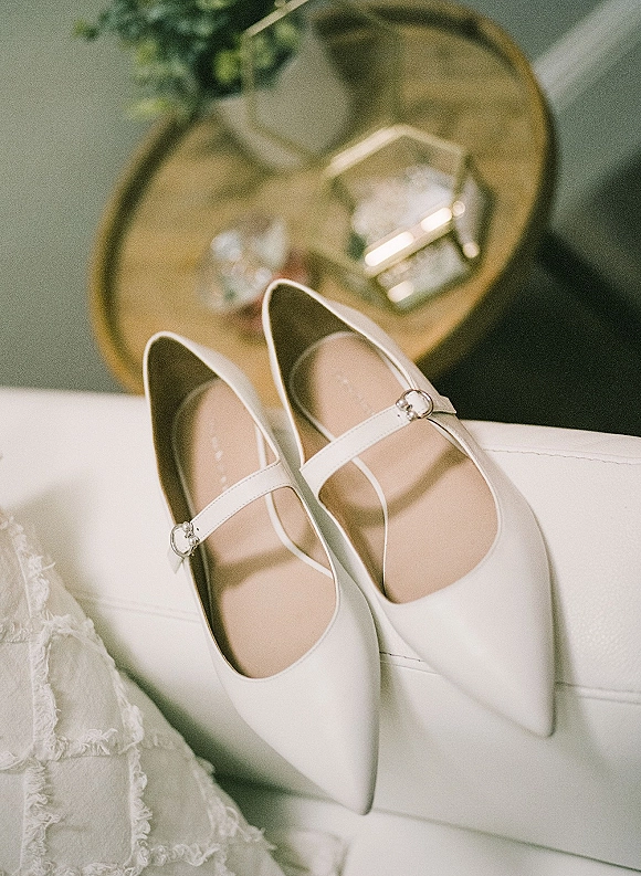 Bridal shoes in classic white wedding heels with mary jane straps and rhinestone buckles on a gold tray beside lace dress on couch