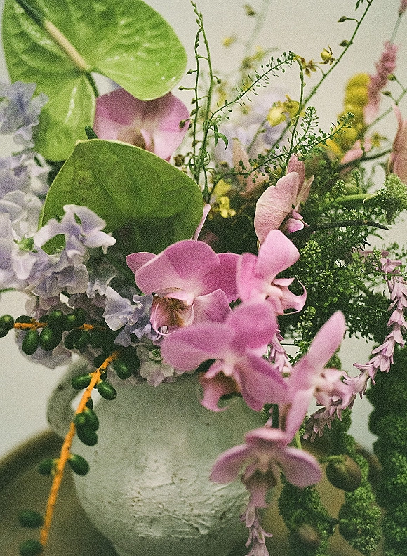 Wedding bouquet with pink orchids and lilac blooms, greenery and seed pods arranged asymmetrically in a ceramic vase against a neutral wall