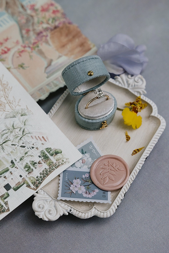 Engagement ring in a blue velvet box with a blush wax seal and ribbon bow on a decorative tray over gray linen with vintage stamps