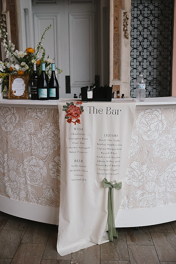Wedding bar signage with a bar menu fabric banner, green ribbon bow, and framed signature drinks sign on a styled bar counter
