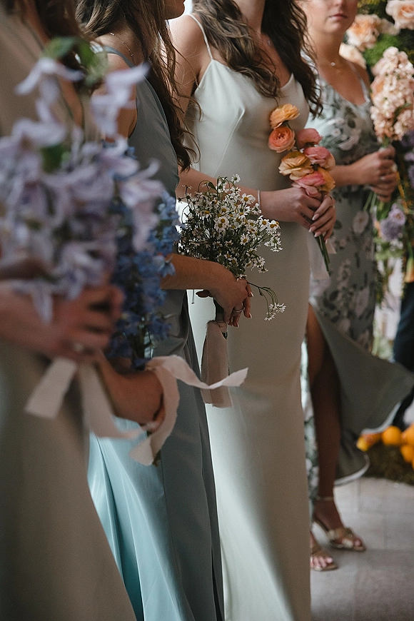 Bridesmaids lineup holding bouquets tied with white ribbon, wearing mix-and-match pastel dresses in soft natural light indoors