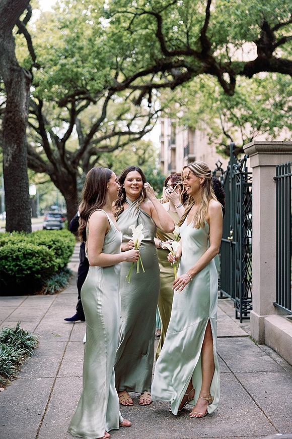 Bridesmaids portrait in sage green bridesmaid dresses, holding white calla lily bouquets on a tree-lined sidewalk by iron fence