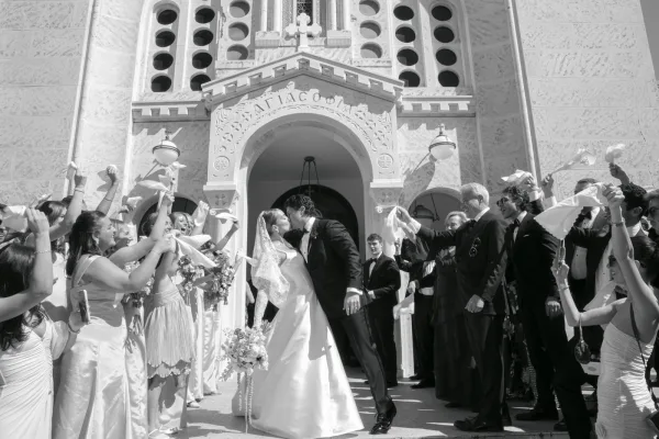 Ceremony exit as the couple shares a wedding exit kiss under the arched church doorway, the bride’s lace veil floating while guests cheer.