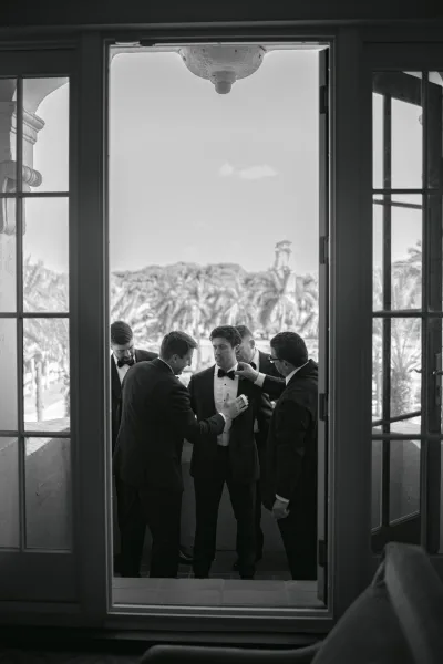 Groom getting ready with groomsmen helping groom adjust his bow tie on a balcony, palm trees and open French doors framing the sky.