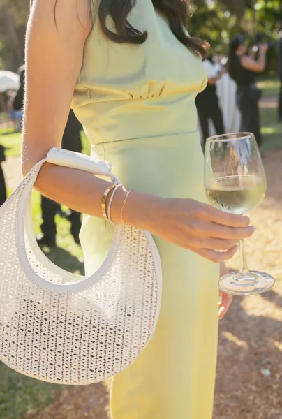 Wedding guest outfit in a sage green guest dress, carrying a woven handbag on a sunlit garden lawn beside a gravel path and trees.