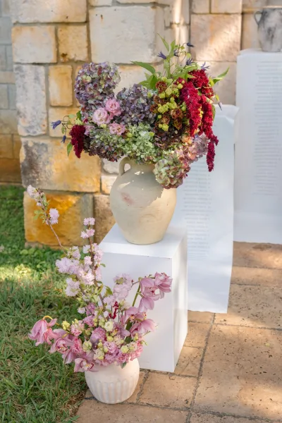 Wedding flower arrangement in ceramic vases, lush blooms set against a stone wall with white backdrop on a grass lawn.