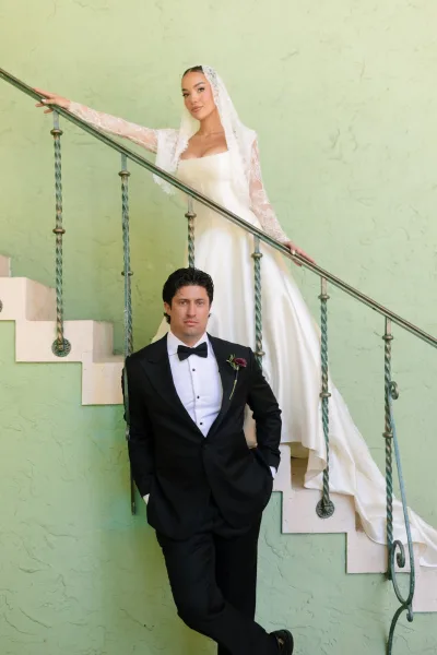 Couple portrait with the bride on a staircase, her lace veil trailing as the groom stands below by a stucco wall and metal railing.