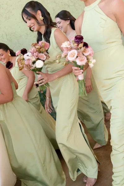 Bridesmaids photo in sage green bridesmaid dresses, holding blush and burgundy bouquets before a textured green wall and wood floor.