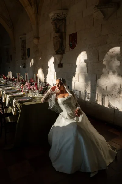 Bridal portrait of a bride leaning on a banquet table, lace veil framing her face, set against an arched stone hallway.