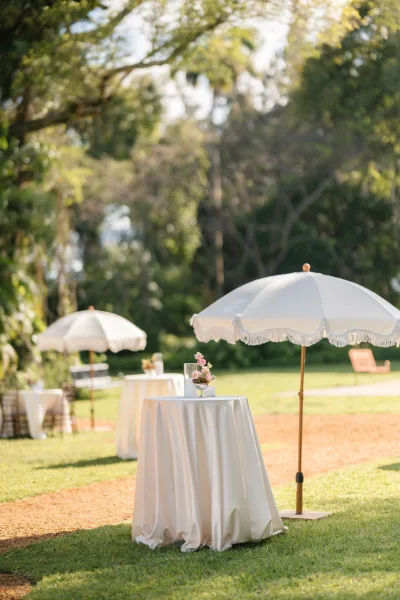 Cocktail hour setup for an outdoor cocktail hour with parasol umbrellas providing shade on a sunlit garden lawn beside a dirt path.