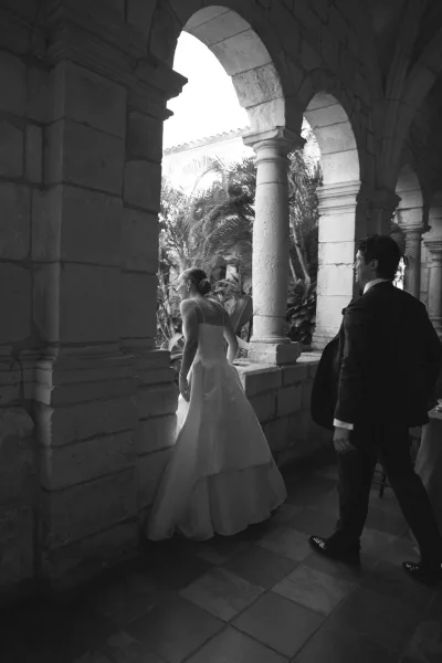 Couple portrait in black and white captures a candid bride and groom, her bridal gown trailing under stone arches and columns on a tiled floor.