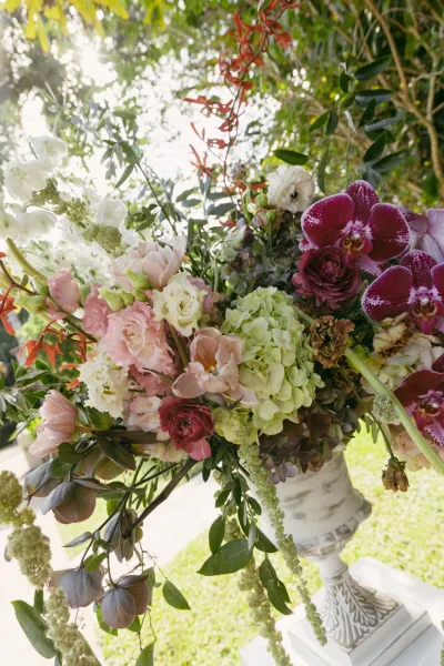 Wedding floral arrangement, a ceremony urn arrangement with orchids, set beside sunlit garden trees and a lawn pathway.