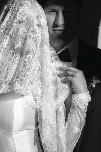 Couple portrait, a black and white wedding portrait where the bride lifts a lace veil and holds the groom's lapel by a stone wall.