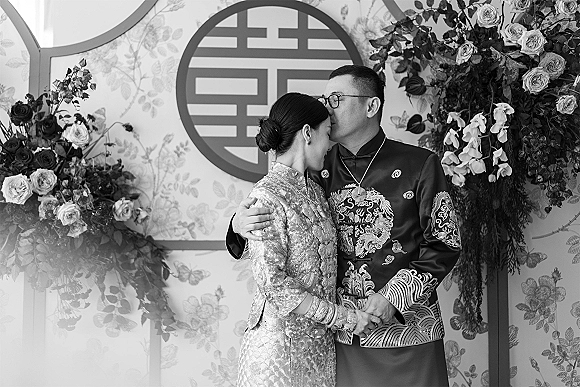 Couple portrait of a traditional wedding couple embracing in embroidered outfits, groom in glasses, with roses, orchids, and a circular backdrop accent