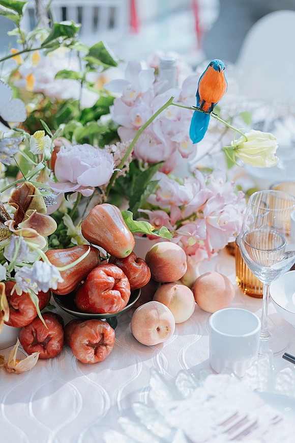 Reception tablescape with wedding table centerpiece of pink orchids, peaches and rose apples, set with wine glasses, plates, amber candle holder in soft daylight