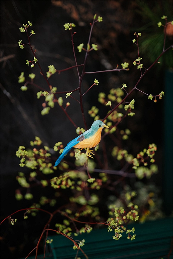 Bird wedding decor with a blue bird cake topper perched among flowering branches and green buds against a dark greenery backdrop