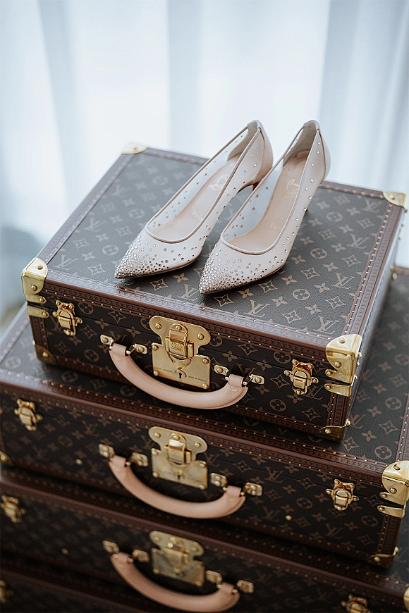 Bridal shoes rhinestone wedding heels perched on stacked luggage trunks with gold hardware, lit by soft window light through sheer curtains