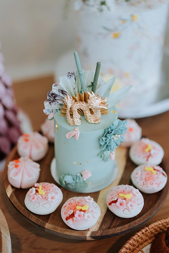 Wedding cake with sage green frosting, gold dragon topper, sugar flowers, taper candles, and iced cookies on a tabletop with vase
