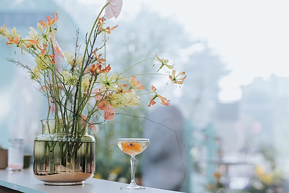 Wedding bar details with a signature cocktail wedding in a champagne coupe, citrus garnish beside a glass vase of wildflowers in window light