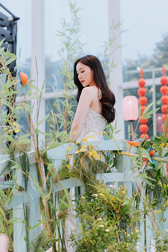 Bridal portrait in a strapless lace wedding dress, bride with long wavy hair looking over shoulder beside greenery and red lanterns indoors