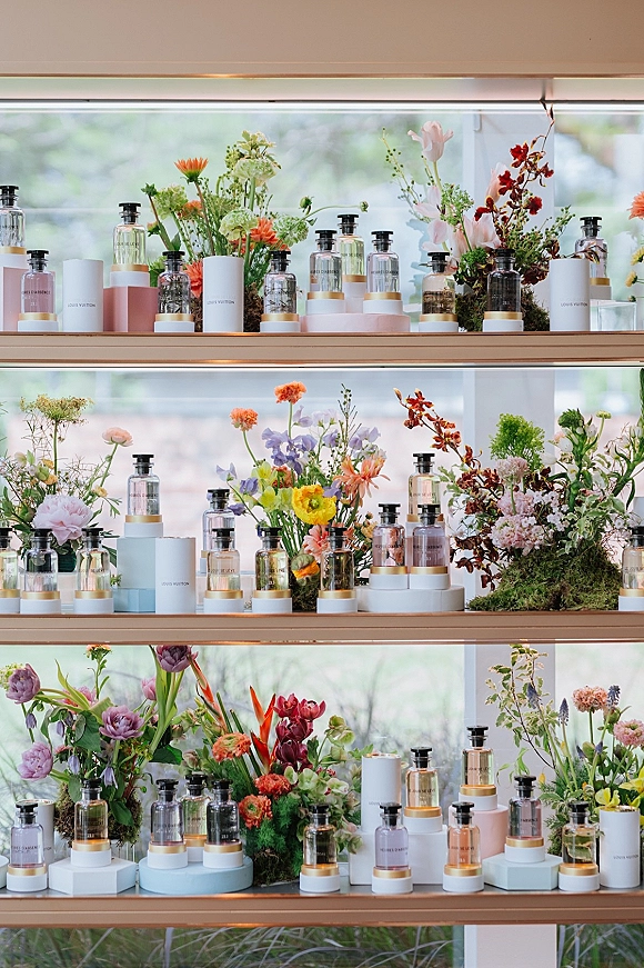 Wedding welcome display with welcome table styling, featuring perfume bottles, pedestal risers, moss, and florals by a window with greenery