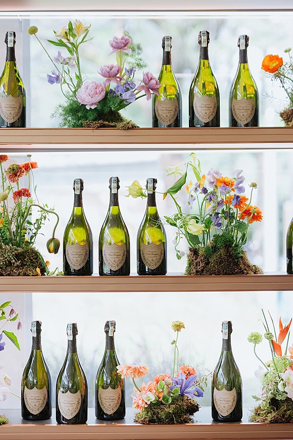 Champagne display for a wedding champagne bar with bottles on shelves, bright florals and moss greenery in a white, window-lit interior