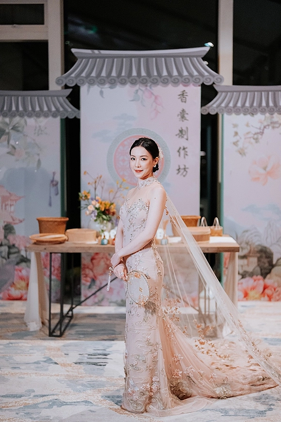 Bridal portrait in a high neck wedding dress with long veil, holding a beaded clutch beside calligraphy signage near large windows indoors