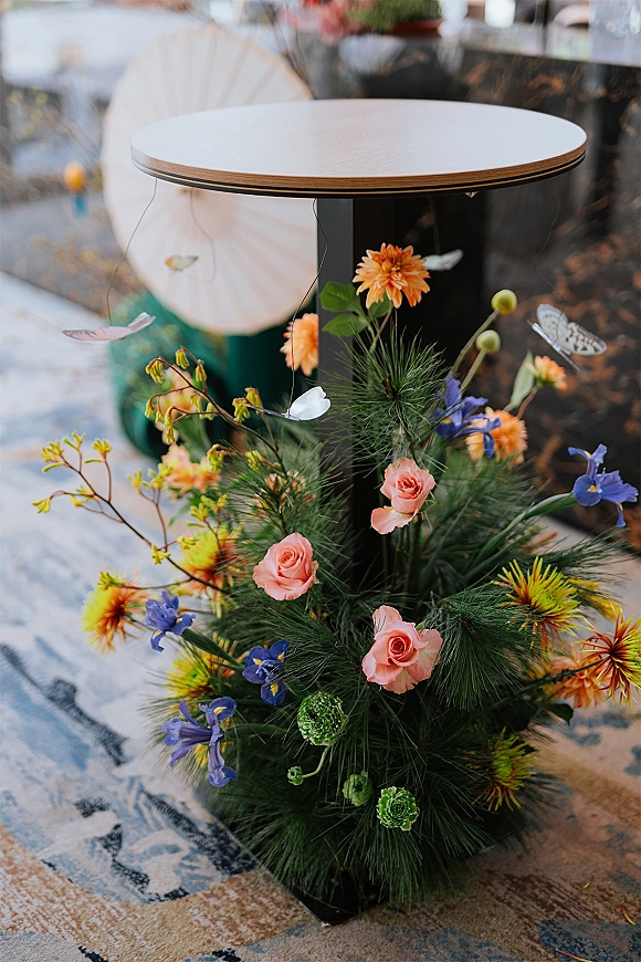 Floral centerpiece wildflower wedding centerpiece with pink roses, orange blooms, purple irises, pine greenery and butterfly accents on a round cocktail table in an indoor lounge setting