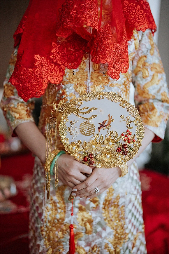 Bridal outfit details with red lace veil and gold embroidered dress, holding a pearl embellished hand fan and wedding ring on a red carpet indoors