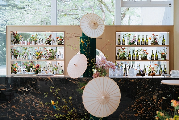 Wedding bar setup with wine bottles and glassware on a black marble bar counter, accented by florals and moss near large windows and trees