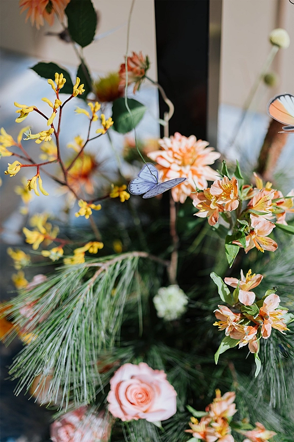 Wedding floral arrangement with orange wedding flowers, pink rose and greenery, accented by a butterfly decoration near a window frame indoors