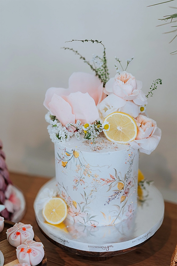 Wedding cake with hand painted floral icing, blush sugar flowers, lemon slices, and chamomile on a stand atop a wood table