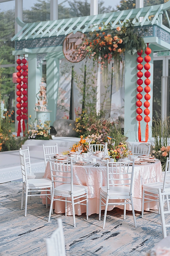 Reception tablescape with a round wedding table setup on blush linens, candlelit florals and fruit centerpiece, plus hanging blooms and red lanterns in a glass-walled garden setting