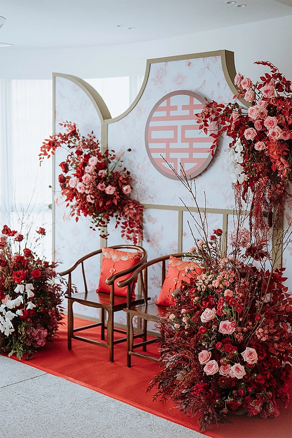 Wedding photo backdrop with lush red rose and orchid installations framing panels and a circular emblem sign, with red carpet and chairs indoors